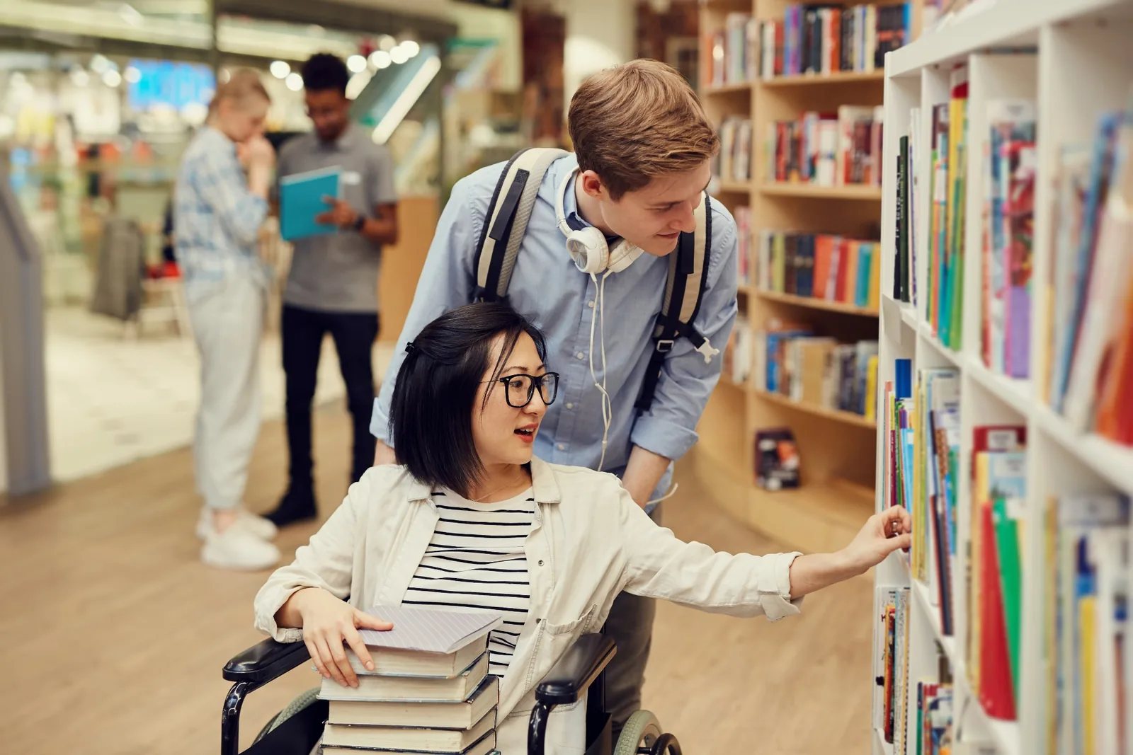 A wheelchair user and another person looking at books on a library shelf