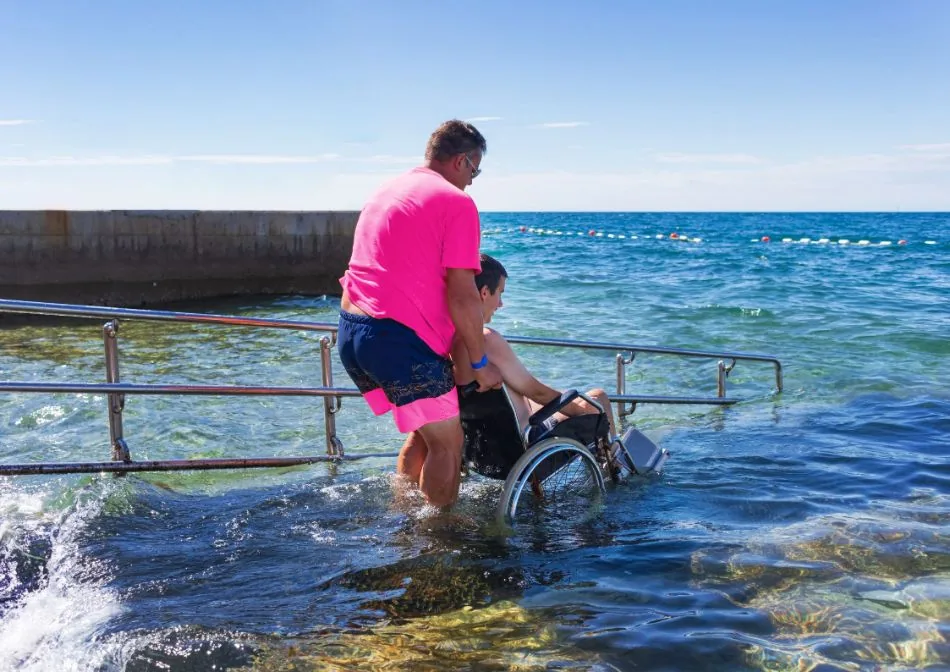 A person assisting a wheelchair user at the edge of the sea