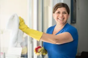 A female cleaner in a blue shirt with yellow gloves cleaning a window with a white towel, while smiling at the camera.