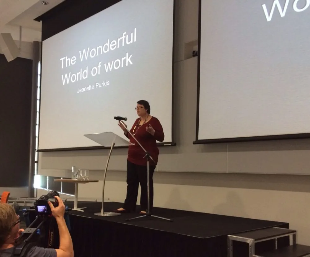 A speaker on stage beside a slide titled The Wonderful World of Work