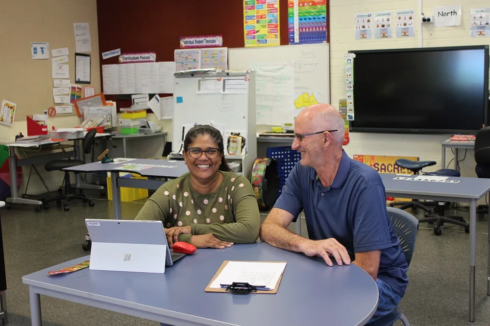 Two adults seated at a classroom table with a tablet