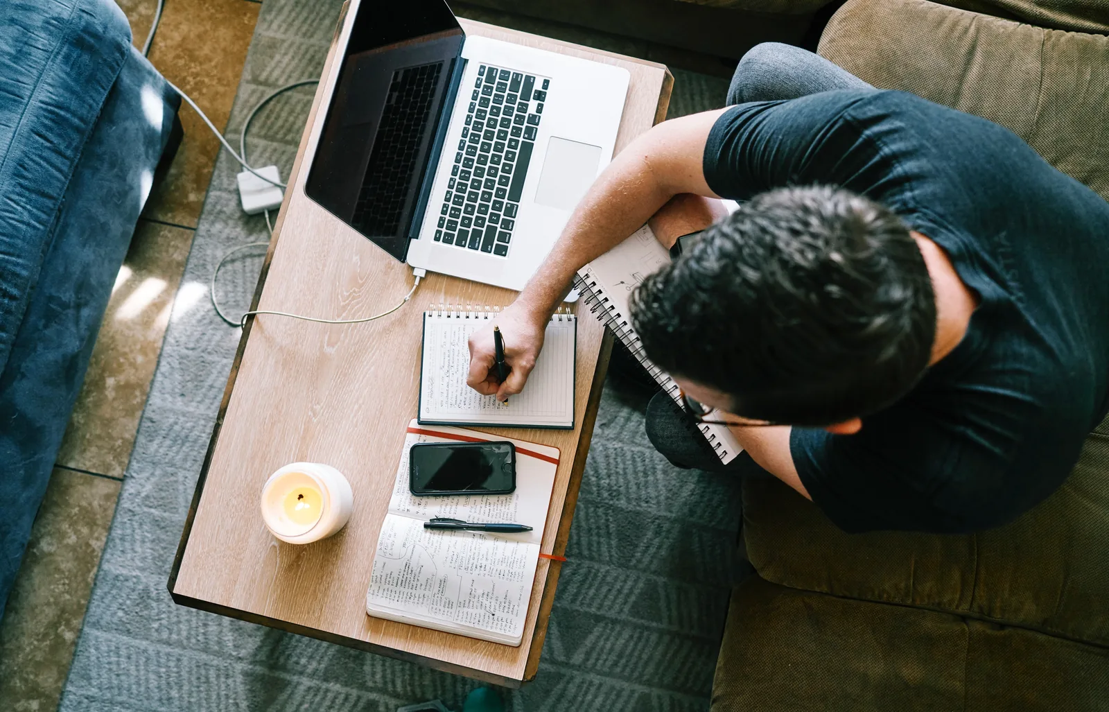 A person writing in a notebook beside a laptop and phone