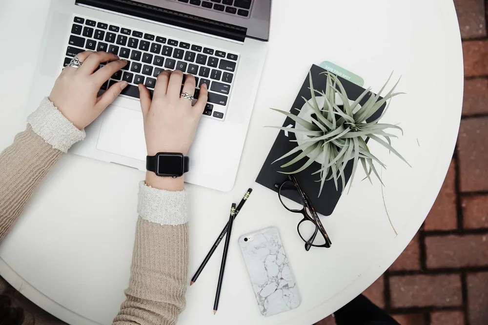 Hands typing on a laptop beside a notebook and plant