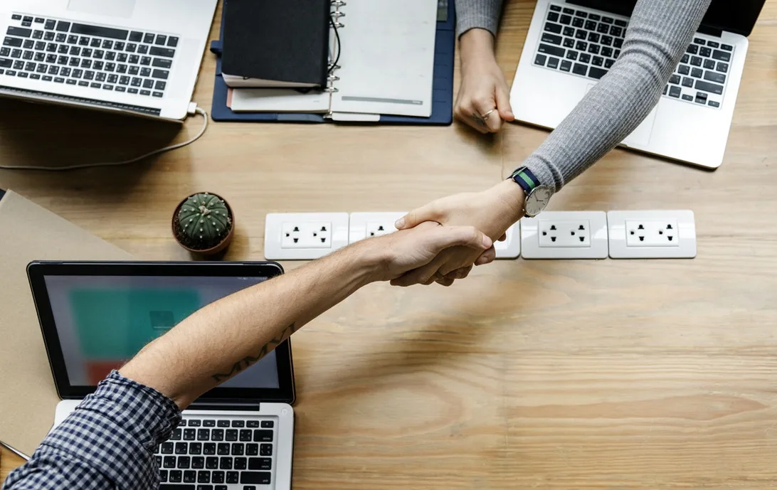 Two people shaking hands across a desk with laptops