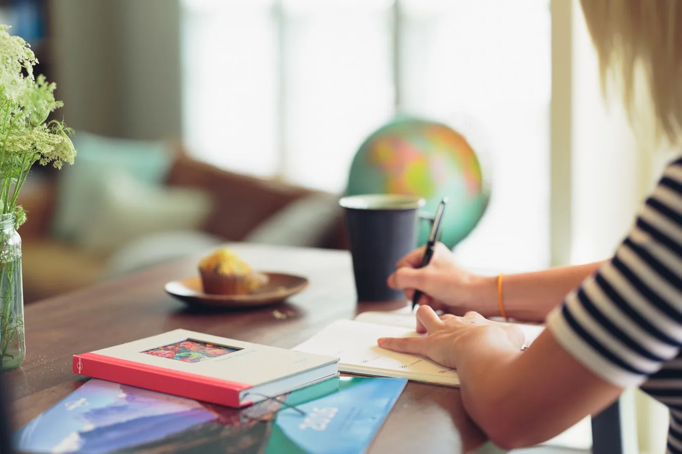 A person writing at a table beside a laptop and books