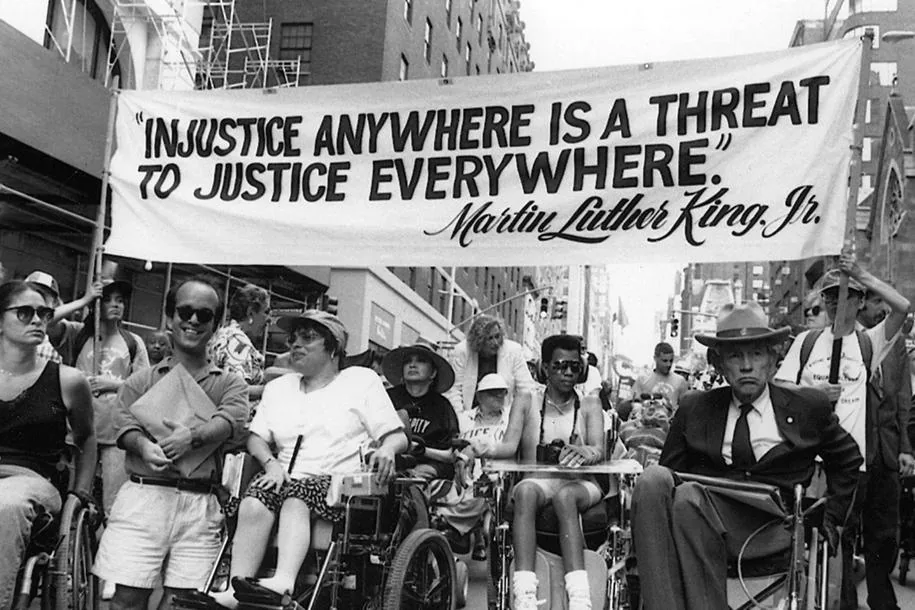A black-and-white photograph of disability rights protesters beneath a banner about justice