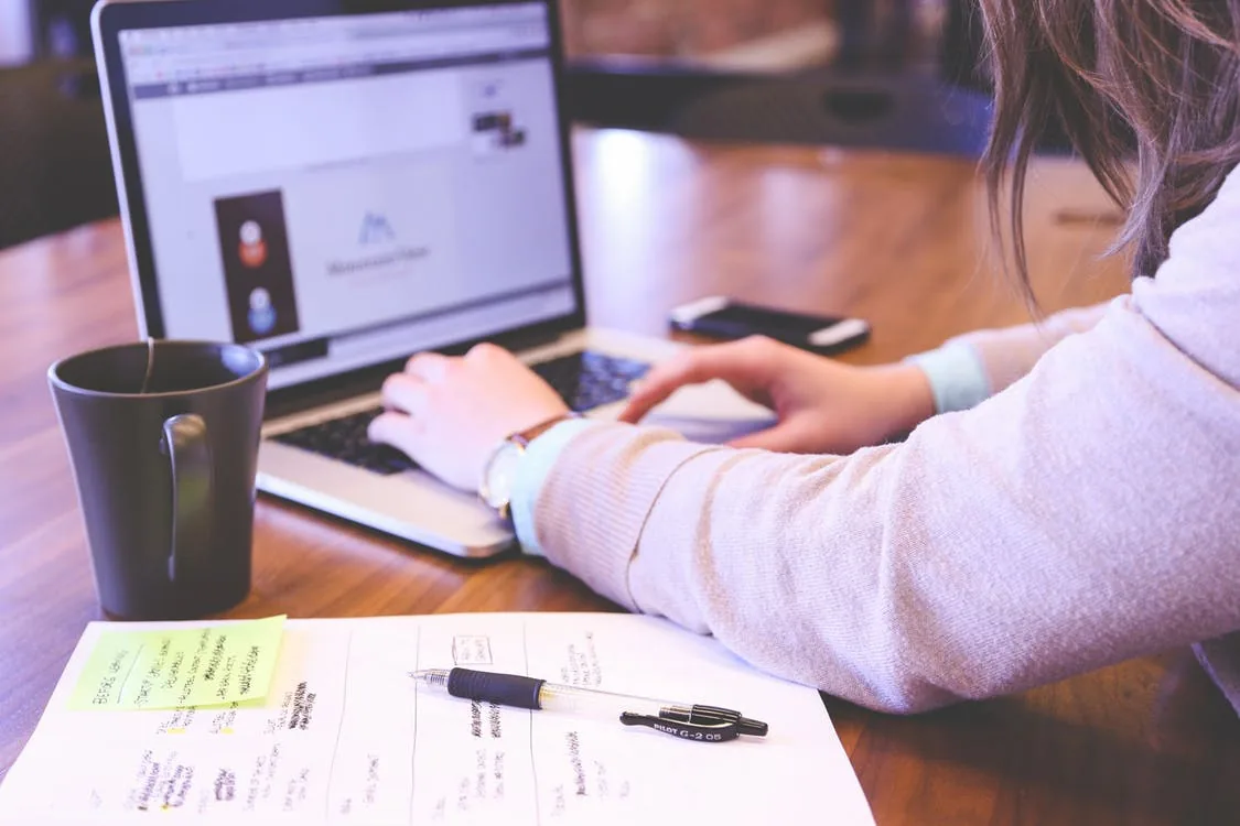 A person working on a laptop beside notes and a cup