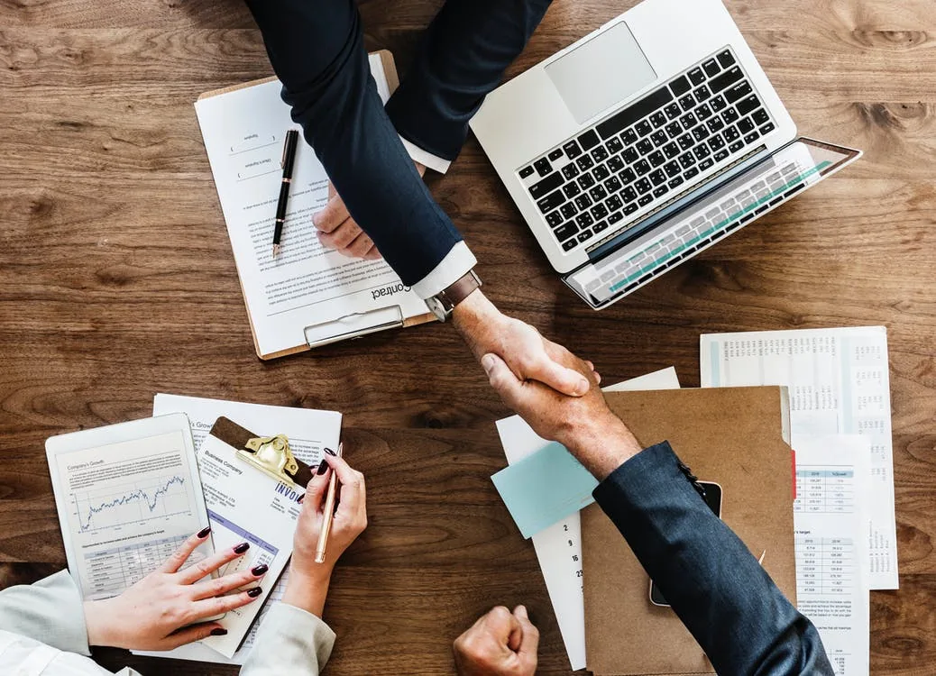 Two people shaking hands over paperwork beside a laptop