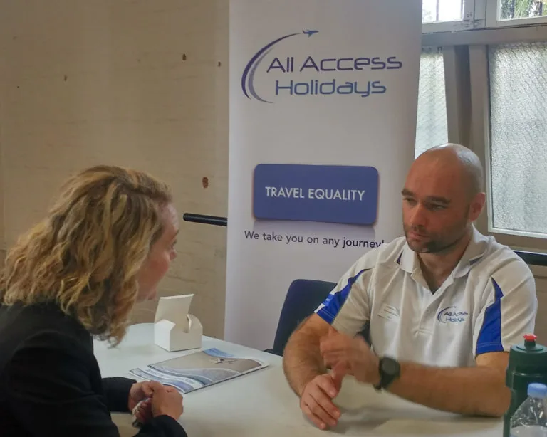 A man in a white polo talking to a woman, in front of a sign that says 'All Access Holidays'.
