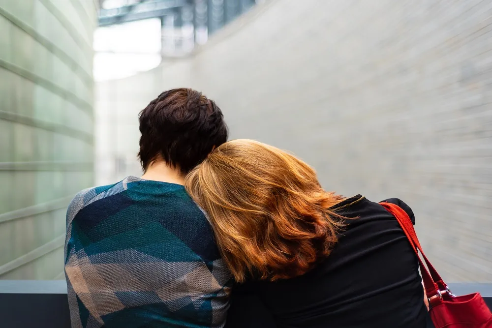 Two people seated together, with one leaning their head on the other’s shoulder