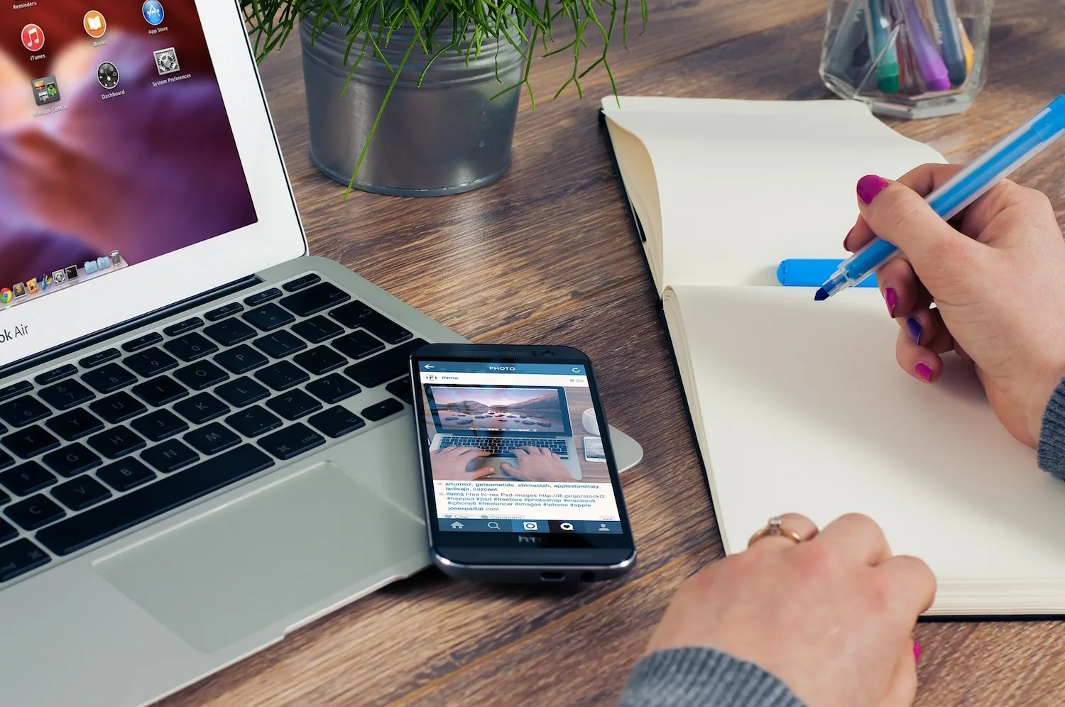 A laptop and phone beside a person writing in a notebook