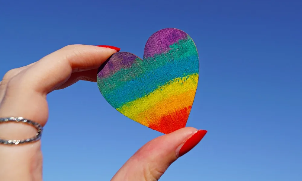 A hand holding a rainbow-coloured heart against the sky
