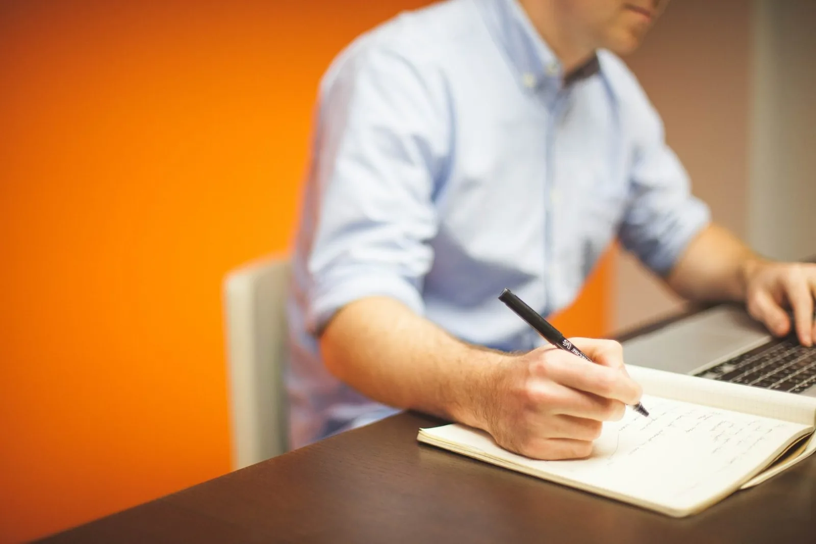 A person writing in a notebook beside a laptop
