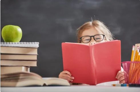 A child wearing glasses reading a red book beside a stack of books
