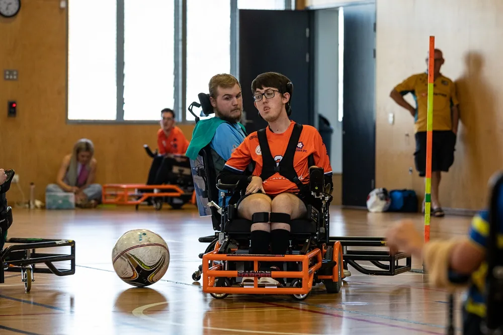 Tom Byrne in an orange powerchair hitting slightly larger soccerball with a guard attached to his chair.