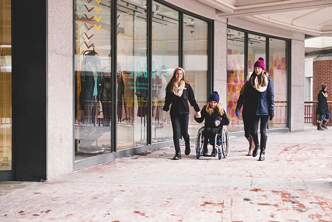 Three people, one in a wheelchair, strolling past shops.
