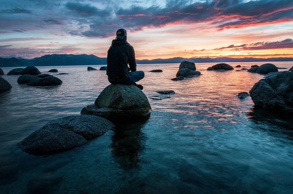 A person seated on a rock beside water at sunset