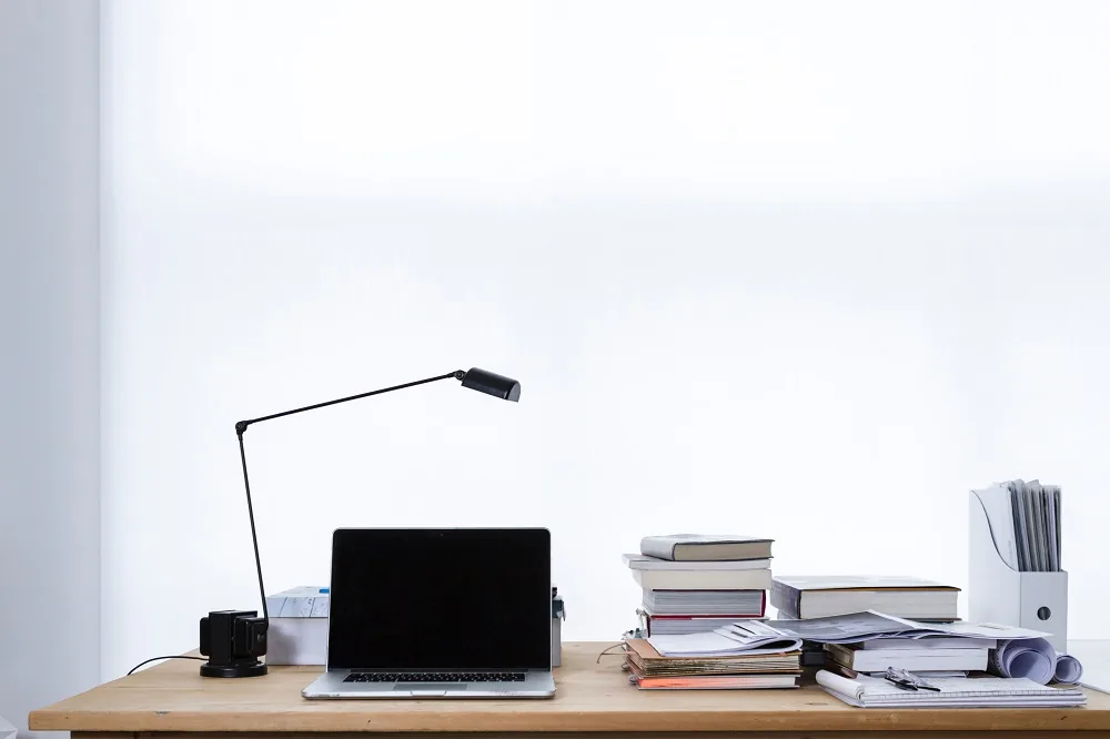 A desk with a laptop, lamp and stacked documents